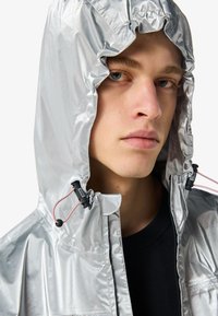 Joven con una chaqueta con capucha plateada brillante y cordones rosas ajustables, mirando ligeramente hacia un lado con una expresión neutral.