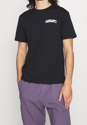 Man wearing black Carhartt t-shirt and purple cargo pants, standing with one hand in pocket against plain white background.