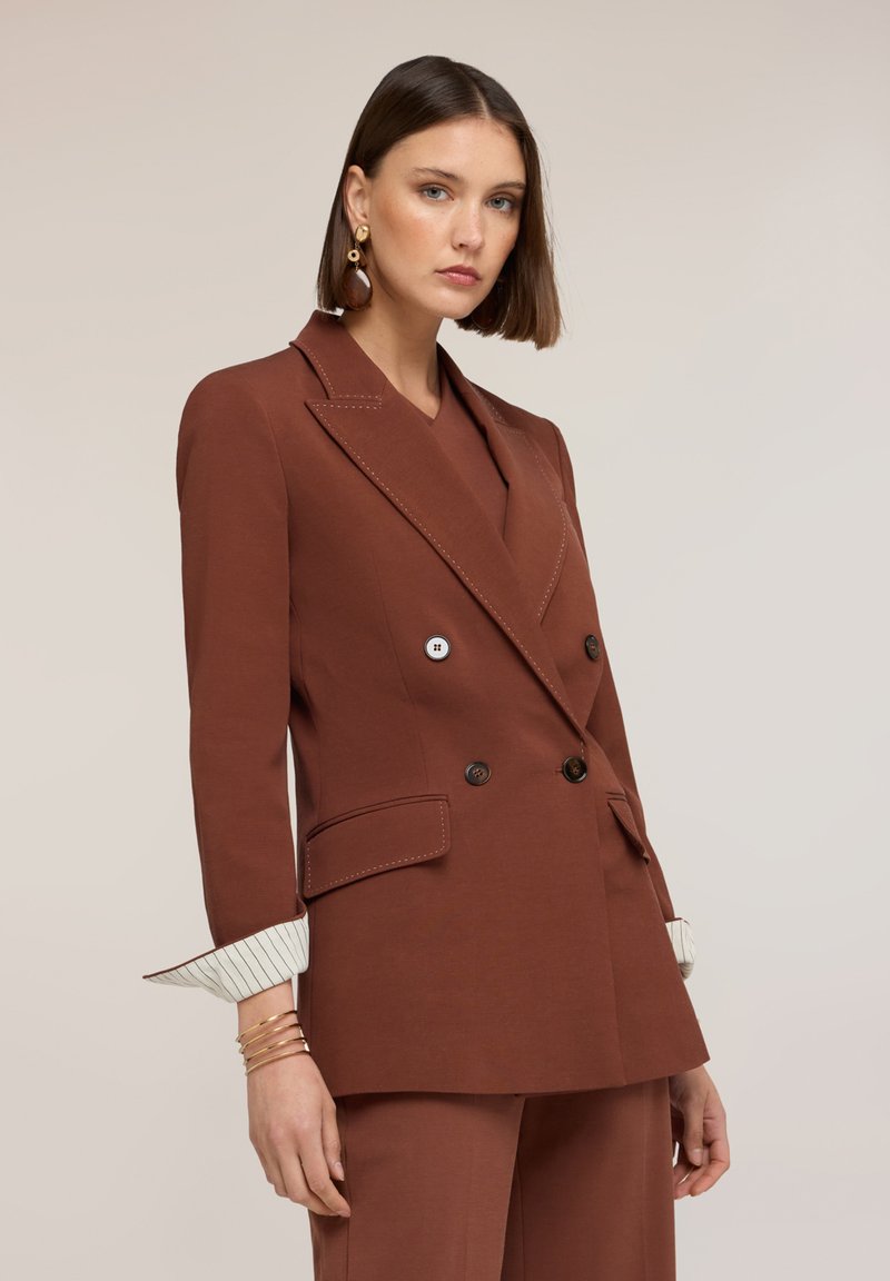 Mujer con cabello corto y castaño que lleva un blazer marrón de doble botonadura y pantalones a juego, complementados con pendientes y pulseras doradas.