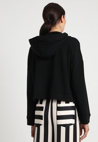 Sudadera corta negra hecha de tela suave con capucha, con hombros caídos y un diseño simple. Combinada con pantalones a rayas.