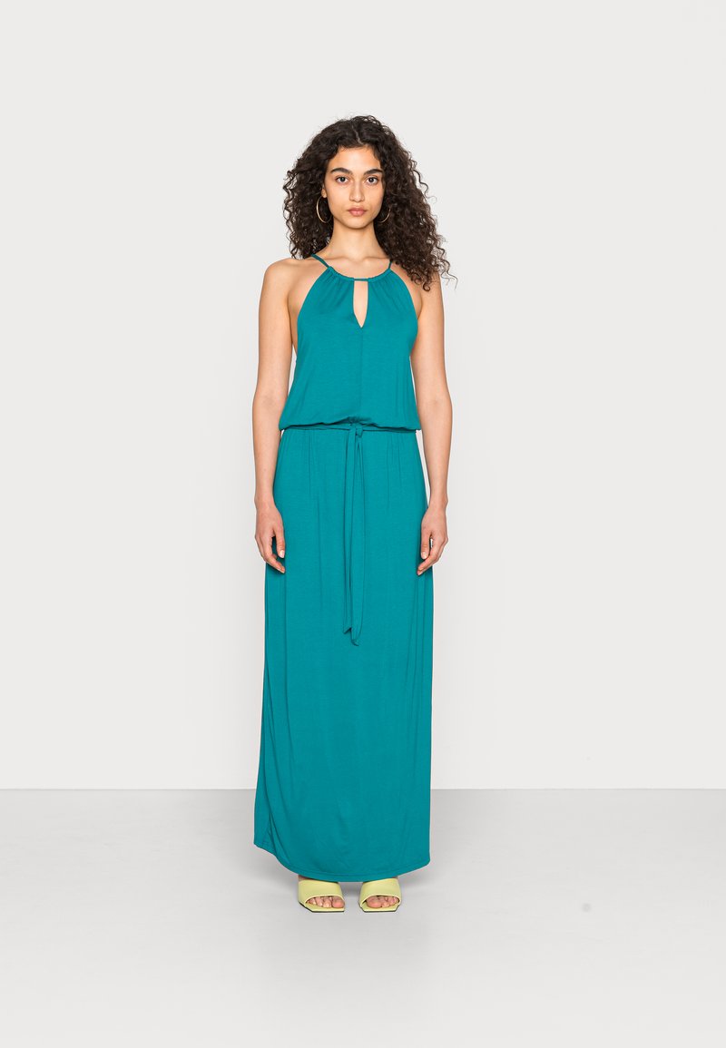 Vestido maxi halter em teal com um decote em forma de chave, cintura cinturada e tecido fluido; usado com sapatos amarelos abertos na frente.