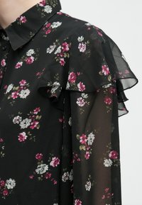 Μαύρη μπλούζα με λουλούδια, διαθέτει διαφανές ύφασμα, ρυαυλοειδή λεπτομέρεια στους ώμους και συνδυασμό ροζ, λευκών και πράσινων λουλουδιών. Κουμπωτός γιακάς.
