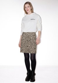 Jeune femme portant un sweat à capuche blanc, une jupe imprimée léopard, des collants noirs et des bottines noires, debout devant un fond blanc uni.