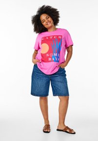 T-shirt graphique rose arborant le texte "CATCH SOME RAYS", associé à un short en jean et des sandales marron. Le t-shirt possède un col rond et une coupe décontractée.