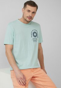 Lichtblauwe katoenen T-shirt met een marineblauw graphic print aan de linkerkant. Gecombineerd met lichtoranje katoenen shorts.