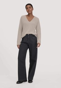 Beige Rippstrickpullover mit V-Ausschnitt, kombiniert mit dunklen, weiten Jeans. Das Model steht aufrecht, trägt schwarze Schuhe und hat einen neutralen Hintergrund.