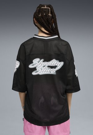 Persona con el cabello trenzado de espaldas, vistiendo una camiseta de malla negra con el texto "Shooting Starz" y pantalones cortos rosas, contra un fondo gris.