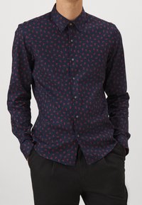 Man som bär en marinblå skjorta med röda geometriska mönster, händerna i fickorna, i kombination med svarta byxor, mot en enkel bakgrund.