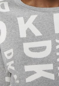 Stříbrný pletený svetr s výraznými bílými textovými vzory, kulatým výstřihem a texturovaným povrchem, který dodává jemný lesk.