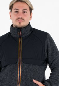 Zwarte en grijze fleecjas met een hoge kraag, ritssluiting aan de voorkant met meerkleurige strepen en duidelijke textuurpanelen.