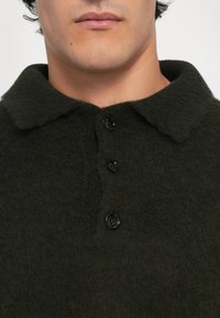 Sötétzöld, textúrált gyapjú pólóing puha gallérral és három fekete gombbal az elején. Közeli nézet a felső testről.