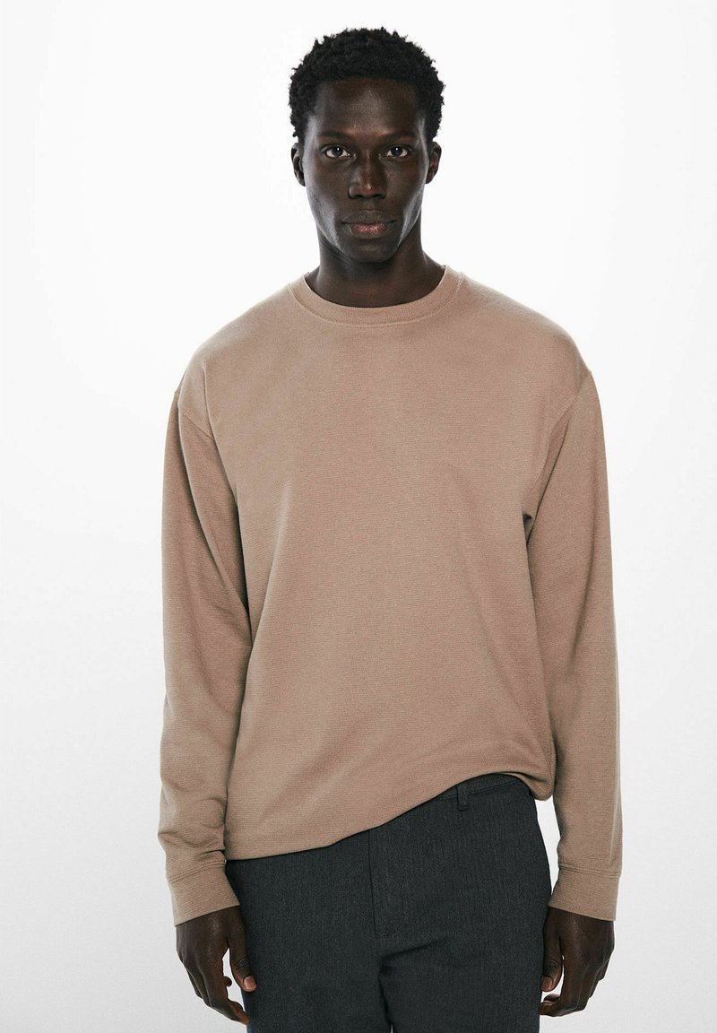 Beige Sweatshirt mit langen Ärmeln aus strukturiertem Stoff, Rundhalsausschnitt, lässige Passform, kombiniert mit dunkelgrauen Hosen, minimalistisches Design.
