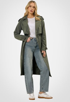 Kvinde iført en olivengrøn trenchcoat, hvid t-shirt, højttaljede blå jeans og hvide sneakers, stående med den ene hånd i lommen.