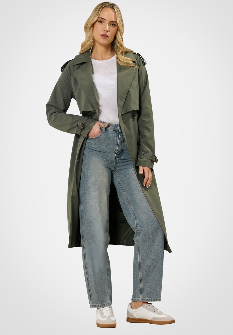 Frau mit olivgrünem Trenchcoat, weißem T-Shirt, hoch taillierter blauer Jeans und weißen Turnschuhen, die mit einer Hand in der Tasche steht.