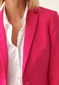 Fuchsiový blazer s texturovaným povrchem, zapínání na jeden knoflík a klopovými límci, oblečený přes bílou košili s knoflíky.