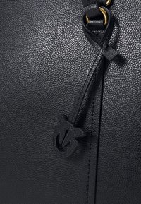 Czarna skórzana torebka o teksturowanym wykończeniu, z wyraźnym akcentem w postaci logo i dekoracyjnym paskiem. Złote okucia widoczne w miejscu połączenia.