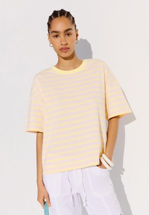 Jeune femme aux cheveux tressés portant un t-shirt ample rayé jaune et rose, un pantalon blanc, des bracelets blancs, posant sur un fond uni.