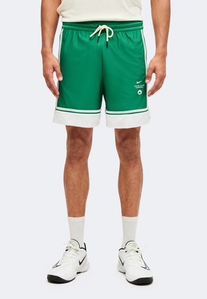 Homme portant un short de basket vert des Boston Celtics avec des bordures blanches, des chaussettes blanches et des baskets blanches Nike, debout devant un fond uni.