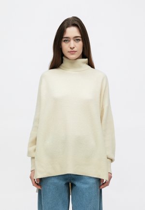 Camisola de gola alta cor creme com um ajuste descontraído, textura canelada e ombros caídos, combinada com jeans azuis claros de perna larga.