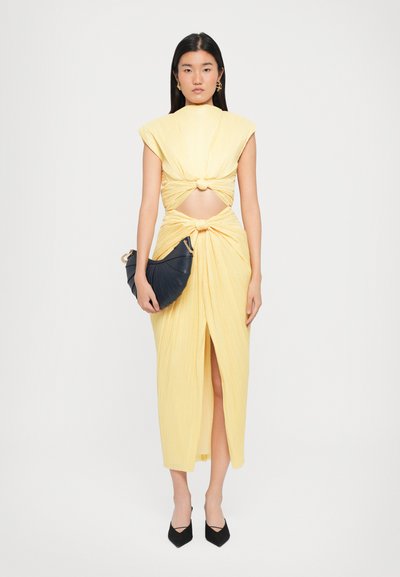 Yellow, sleeveless dress with a knotted design, cut-outs, and a mid-length hem. Complemented by a curved black bag and pointed shoes.