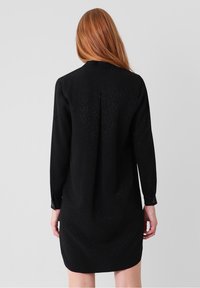Femme aux longs cheveux roux portant une robe noire à manches longues, debout de dos sur un fond gris clair uni.