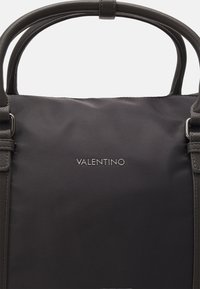 Sac en tissu noir avec une texture lisse, des poignées doubles, et le logo argenté "VALENTINO" sur le devant, avec des accents en cuir sur les côtés.