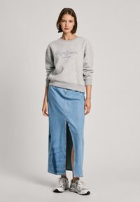 Sudadera gris con mangas ajustadas y logo delantero, combinada con una larga falda de mezclilla azul con una abertura frontal, y zapatillas deportivas grises.