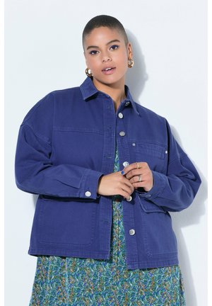 Person med kort hår iført gyldne creoler, en blå knappet jakke over en blomsterkjole, poserer mod en lys baggrund.