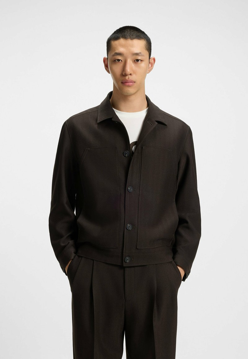 Dark brown, short jacket with a collared neckline, front pockets, and button closure. Made from textured fabric with a tailored fit.