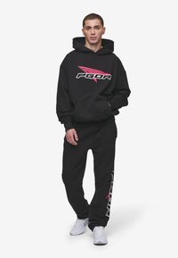 Felpa nera con logo PGDR in rosa e bianco, abbinata a pantaloni della tuta neri coordinati. Il tessuto sembra morbido, con una vestibilità comoda e tasche.