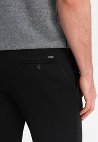 Schwarze Hose aus strukturiertem Stoff, mit einer hinteren Tasche mit Knopf und einem kleinen metallischen Akzent. Das Oberteil ist ein graues, gemustertes Hemd.