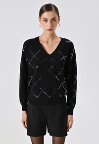 Maglione nero con scollo a V, caratterizzato da un motivo a diamante in paillettes argento. Texture morbida, vestibilità ampia, con polsini e orlo a coste. Abbinato a pantaloncini neri.