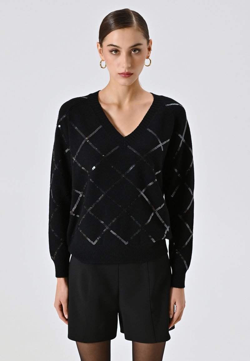 Maglione nero con scollo a V, caratterizzato da un motivo a diamante in paillettes argento. Texture morbida, vestibilità ampia, con polsini e orlo a coste. Abbinato a pantaloncini neri.
