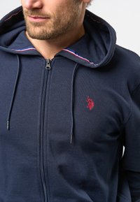 Mörkblå zip-up hoodie i bomullsblandning med kontrasterande randig kant på huvan och liten röd logga på bröstet. Texturerad yta.
