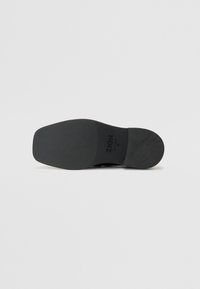 Suela de zapato negra con una superficie de goma texturizada, que presenta una punta redondeada y un diseño plano. Logo visible en la suela.