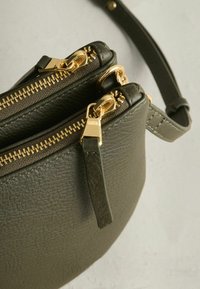 Olivgrüne, strukturierte Lederhandtasche mit zwei goldfarbenen Reißverschlüssen, goldfarbenen Beschlägen und einem abnehmbaren Schultergurt.