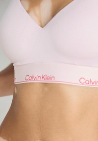 Roze bralette met een V-hals ontwerp, elastische band met het witte en rode "Calvin Klein" logo. Soepele textuur en een aansluitende pasvorm.