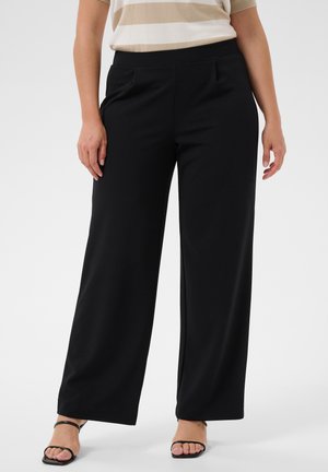 Femme portant un pantalon noir à jambes larges et des talons noirs ouverts, debout devant un fond uni.