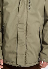 Veste isolante vert olive avec fermeture éclair et bouton-pression. Présente des poches latérales, un tissu texturé et un logo distinctif à l'avant.