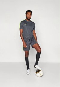 Man in donkergrijze sportkleding met neonaccenten staat met één voet op een wit-zwart voetbalt tegen een effen achtergrond.