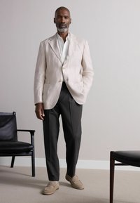 Helle beige Leinenblazer mit zwei Knöpfen, kombiniert mit dunkel gestreiften Hosen und neutralen Slippern. Minimalistischer schwarzer Stuhl im Hintergrund.