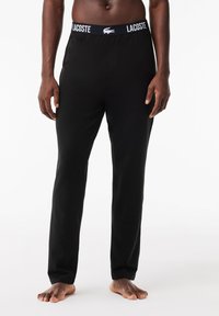 Schwarze Lounge-Hosen aus weicher Baumwolle mit einem elastischen Bund, versehen mit einem weißen "LACOSTE"-Logo und Krokodil-Emblem.