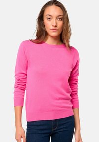Pull-over rose en tissu doux, avec des manches longues, un col rond et des poignets et un ourlet côtelés. Porté avec un jean foncé.
