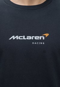 Μαύρο πουκάμισο με λευκό κείμενο "McLaren" και πορτοκαλί λογότυπο πάνω από μικρότερο λευκό κείμενο "Racing" στην περιοχή του στήθους.