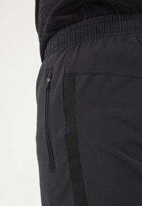 Svarta sportshorts med elastiskt midjeband, med en ficka med blixtlås och texturerade sidopaneler. Tyget verkar slätt och hållbart.