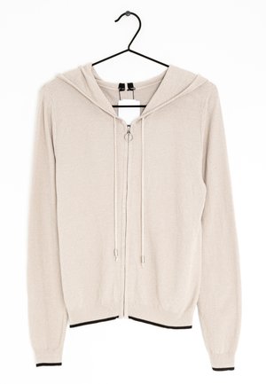 Sudadera con capucha beige de tejido de punto y cremallera, con cordones, colgada en una percha negra, con tela texturizada y puños y dobladillo acanalados con ribete negro.