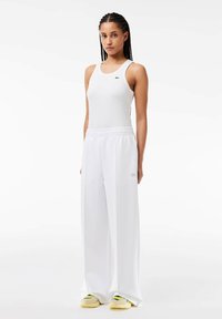 Un top fără mâneci alb cu nervuri și pantaloni albi lejeri, având un logo subtil pe pantaloni. Modelul poartă adidași deschiși cu accente verzi.