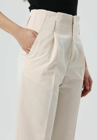 Beige, hoch taillierte Hose mit seitlichen Taschen, plissiertem Vorderteil und glatter Stoffstruktur. Sichtbare Nahtakzente entlang des Bunds.