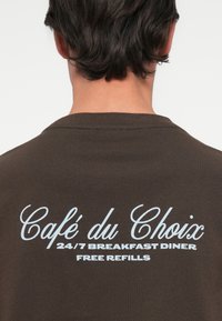 Καφέ καφέ κοντομάνικο μπλουζάκι με λευκό κείμενο στην πλάτη: "Café du Choix 24/7 BREAKFAST DINER ΔΩΡΕΑΝ ΕΠΙΣΚΟΠΗΣΕΙΣ." Λείος υφασμάτινος.
