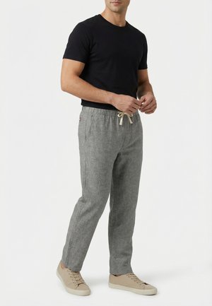 Uomo con una maglietta nera a maniche corte, pantaloni grigi con coulisse e sneakers beige, in piedi davanti a uno sfondo bianco.